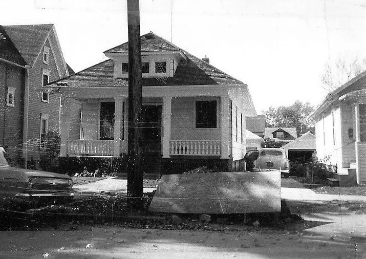 Black-and-white photo of a single-story bungalow with a front porch and central steps, a utility pole in front, vintage cars parked on the street and in the driveway leading to a detached garage.