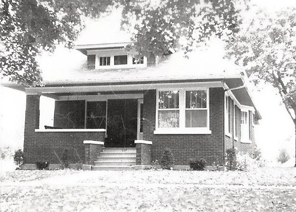 Black-and-white photo of a one-story brick bungalow with a covered front porch, central steps, dormer window, and trees framing the yard.