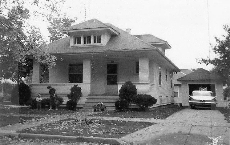 Black-and-white photo of a 1950s-era bungalow with a wide front porch, shrubs, two children near the steps, and a vintage car parked in the driveway by a detached garage.