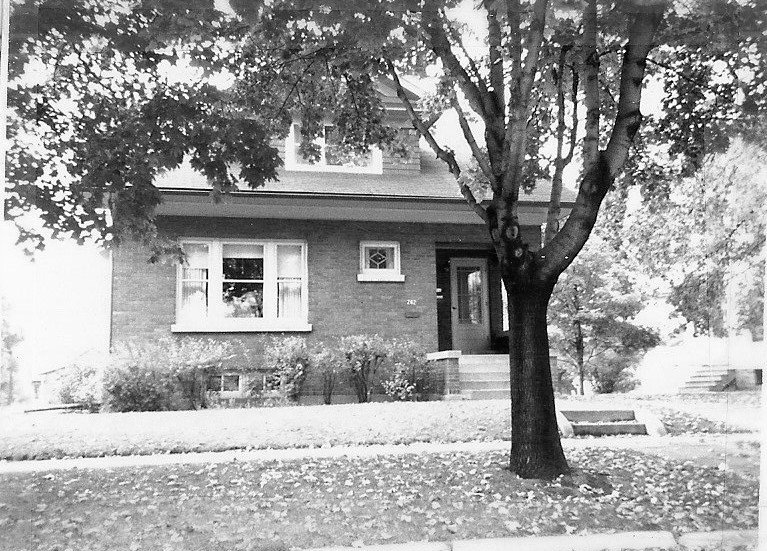 Black-and-white photo of a two-story brick house with a front porch entrance, windows, a large tree in the front yard, and fallen leaves on the lawn.