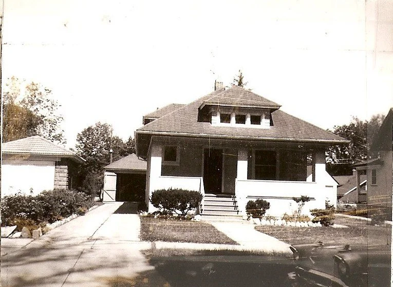 Black-and-white photo of a single-story bungalow with front porch, steps, shrubs, driveway leading to a detached garage, and a partial vintage car in the foreground.