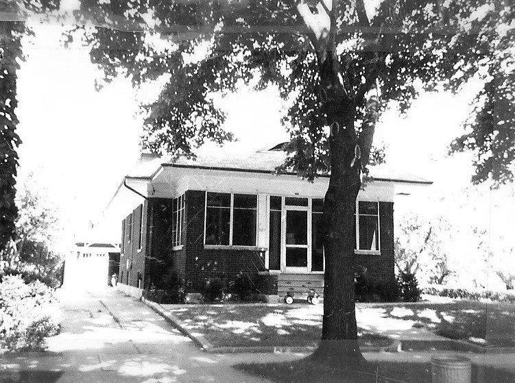 Black-and-white photo of a single-story brick bungalow with an enclosed front porch, large tree in the front yard, driveway leading to a detached garage.