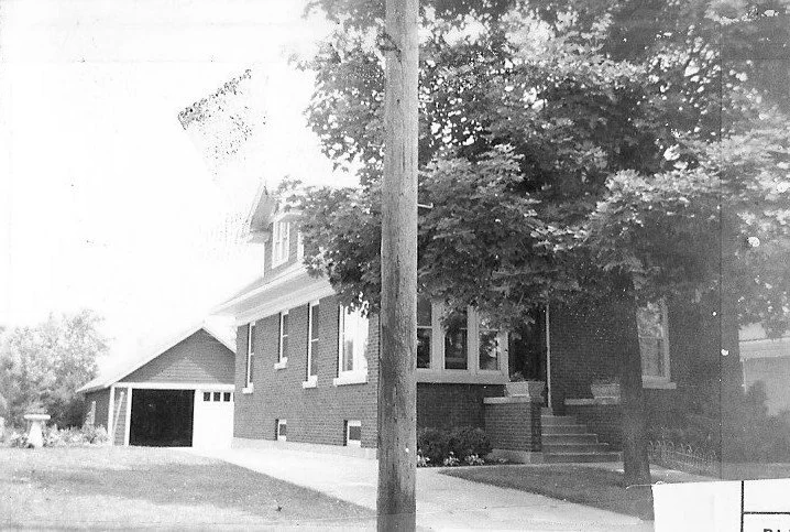 Black-and-white photo of a brick bungalow with a detached garage, large front tree, sidewalk and a utility pole in the foreground.