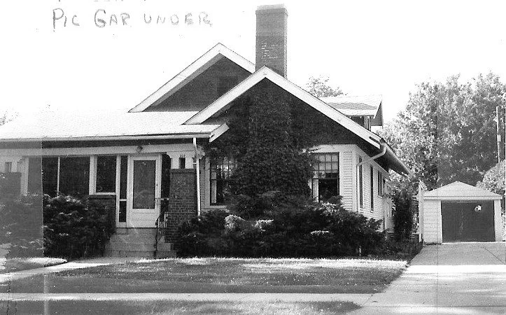 Black-and-white photo of a single-story bungalow with a front porch, central chimney, shrubs, and a detached garage at the end of the driveway.