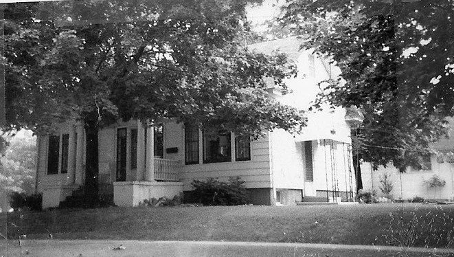 Black-and-white photo of a single-family house with a front porch, large overhanging trees, and a grassy lawn.