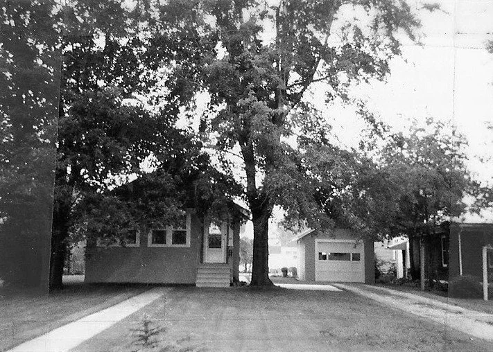 Black-and-white photo of a small single-story house with a large tree in front and a driveway leading to a detached single-car garage.