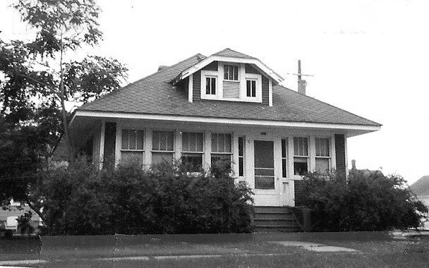 Black-and-white photo of a single-story bungalow with a front enclosed porch, dormer window, and shrubs lining the walkway.