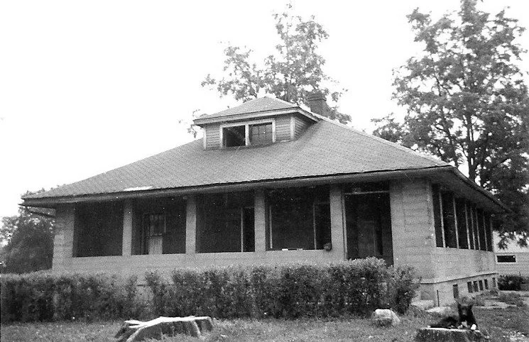 Black-and-white photo of a one-story bungalow with a wide screened front porch, low hipped roof with a small dormer, and shrubs and trees around it.
