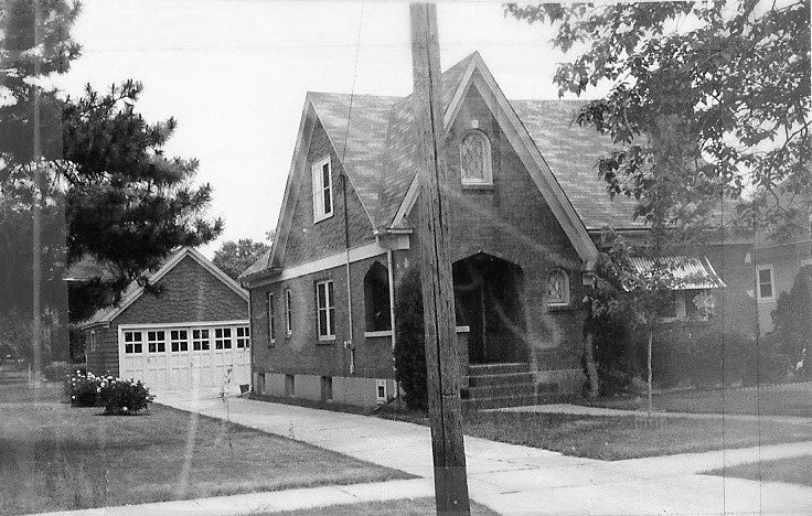 Black-and-white photo of a brick gabled house with a front porch, detached garage, driveway, lawn, and a utility pole in the foreground.