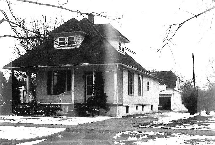 Black-and-white photo of a two-story house with a covered front porch, dormer windows, driveway leading to a detached garage, and patches of snow on the ground.