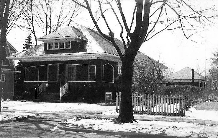 Black-and-white photo of a bungalow-style house with a covered front porch, bare tree in the foreground and a small picket fence with light snow on the ground.