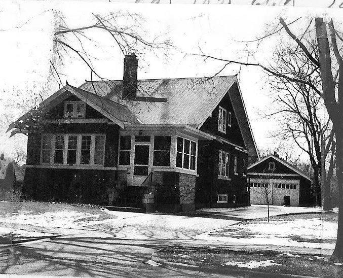 Black-and-white photo of a two-story bungalow with a front porch and stone foundation, a detached garage to the right, bare trees and light snow on the ground.