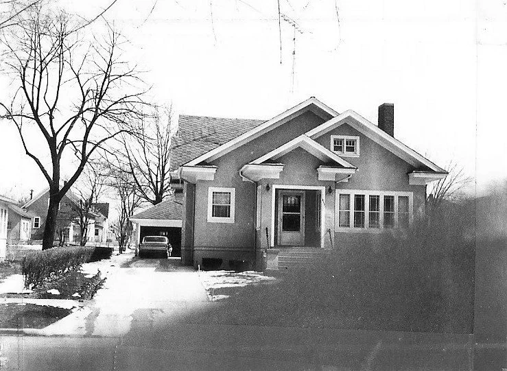 Black-and-white photo of a single-story bungalow with a front porch, detached garage and car in the driveway, bare trees and patches of snow in the yard.