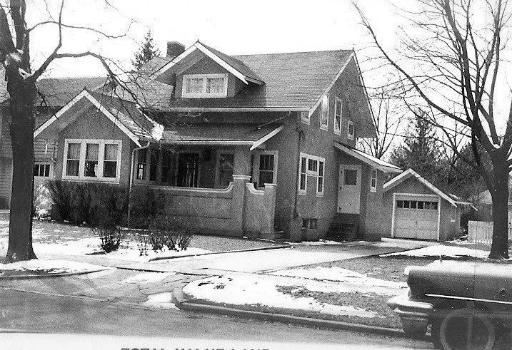 Black-and-white photo of a two-story house with a covered front porch, detached garage to the right, leafless trees and patches of snow on the lawn; a car bumper is visible in the foreground.