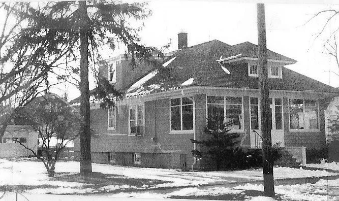 Black-and-white photo of a two-story bungalow with an enclosed front porch, snow on the ground and bare trees in the yard.