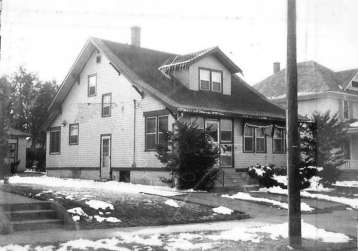 Black-and-white photo of a two-story gabled house with a covered porch, snow patches on the lawn, and a utility pole in the foreground.
