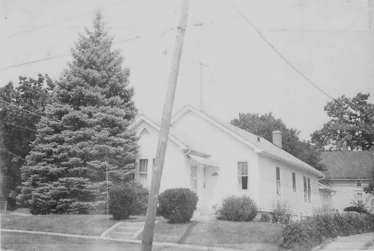 Black-and-white photo of a single-story clapboard house with a front porch, large evergreen tree to the left, shrubs and a leaning utility pole in the yard.