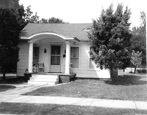 Black-and-white photo of a small white bungalow with a columned front porch, two lawn chairs, and a large tree on the right.