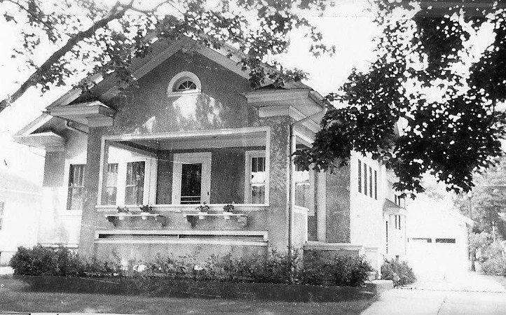 Black-and-white photo of a two-story bungalow with an enclosed front porch, flower boxes under the windows, a tree in the foreground and a driveway leading to a detached garage.