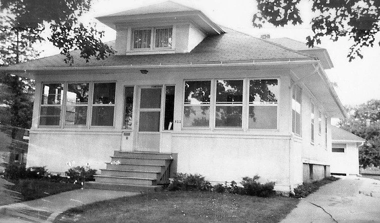 Black-and-white photo of a single-story bungalow with an enclosed front porch, central steps, and a driveway leading past the right side.