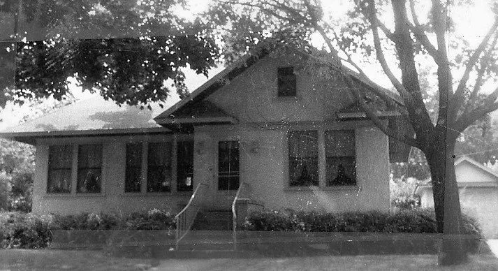 Black-and-white photo of a single-story bungalow with a small front porch, steps and metal railings, flanked by trees.