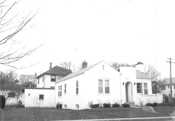 Black-and-white photo of a single-story white stucco house with an arched front entrance, small lawn, and neighboring homes in the background.