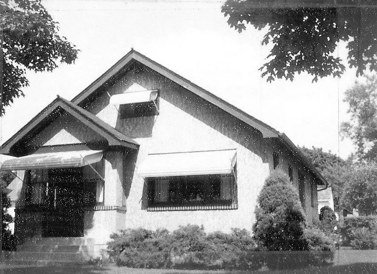 Black-and-white photo of a single-story bungalow with window awnings, front steps, and shrubs in the yard.