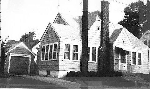 Black-and-white photo of a small single-story house with a tall brick chimney, nearby detached garage, and a utility pole in the foreground.