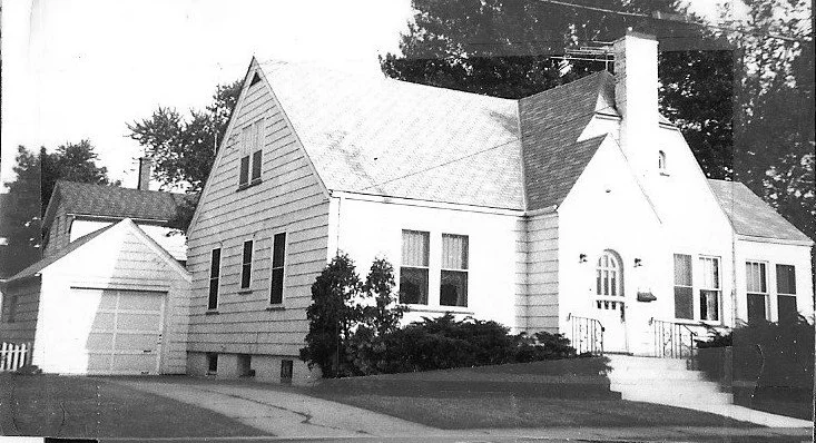 Black-and-white photo of a mid-century single-family house with steep gabled roof, arched front door, front steps, shrubs, and a detached garage.