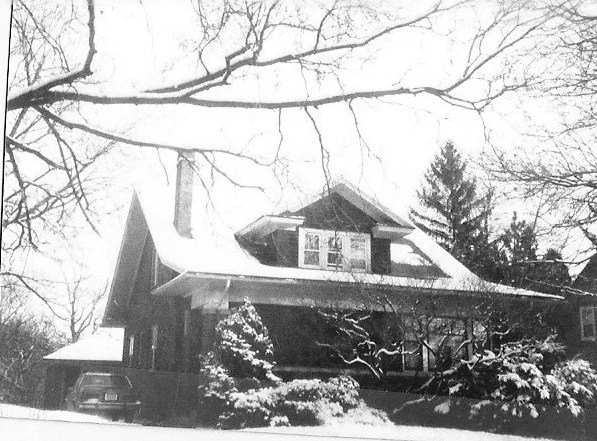 Snow-covered two-story house with a front porch and dormer window, bare tree branches overhead and evergreens in the yard.