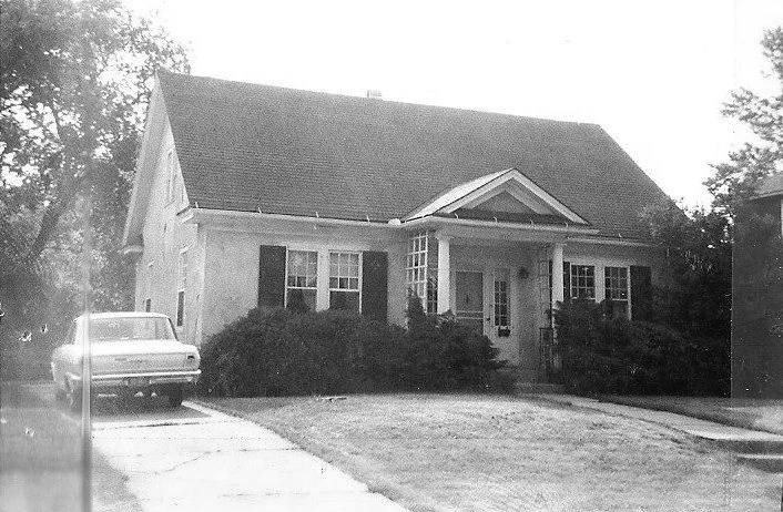 Black-and-white photo of a one-story suburban house with a steep pitched roof, small front porch flanked by columns, shrubbery, and a car parked in the driveway.