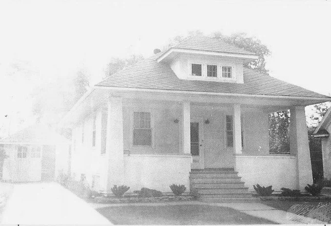 Black-and-white photo of a single-story bungalow with a covered front porch, wide steps and square columns, a dormer window on the roof, and a detached garage to the left.