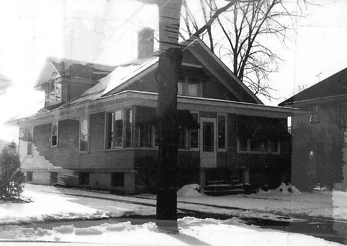 Black-and-white photo of a snow-covered early-20th-century bungalow with a front porch, striped awning, and a bare tree in front.