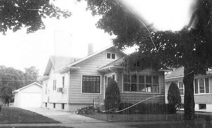 Black-and-white photo of a single-story bungalow with a screened front porch, driveway leading to a detached garage, and large trees partially shading the house.