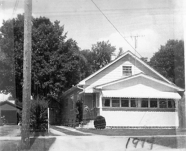 Black-and-white photo of a single-story house with an enclosed front porch, driveway, large trees behind it, and a telephone pole in the foreground.