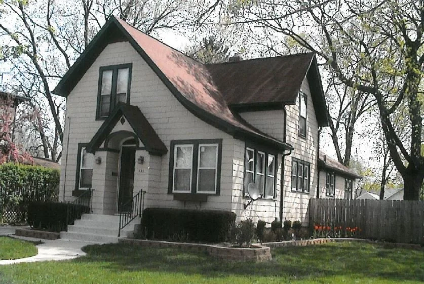 A two-story cream-colored house with steep gabled roof, front steps, lawn, and surrounding trees.