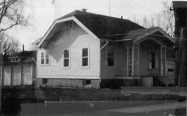 Black-and-white photo of a small single-story wood bungalow with front porch and attached garage, leafless trees in yard.