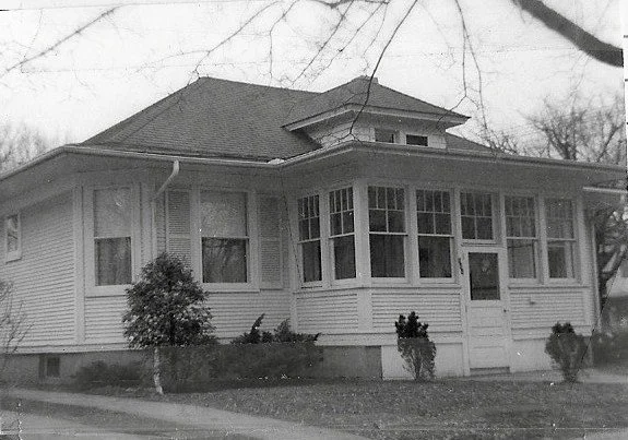 Black-and-white photo of a single-story wood-frame bungalow with an enclosed front porch, dormer roof, and small shrubs in front.