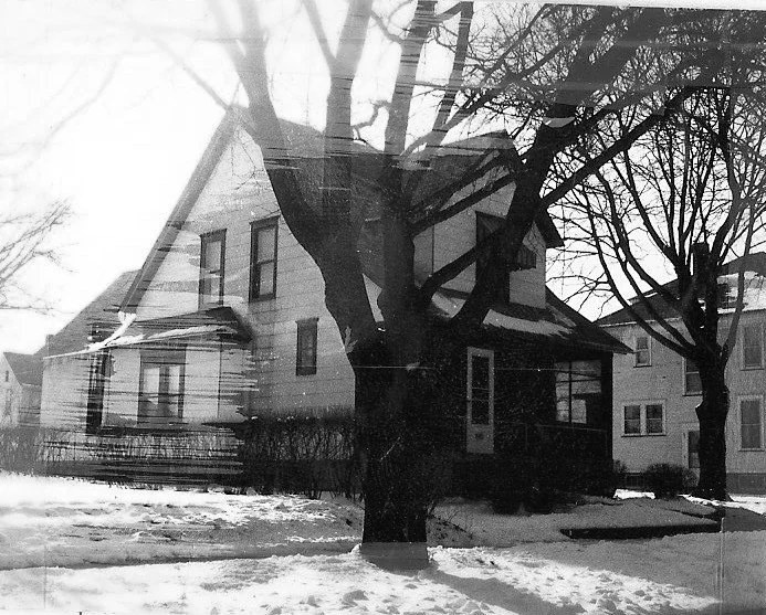 Black-and-white photo of a two-story house in winter with bare trees in front and snow-covered lawn.