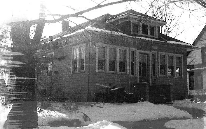 Black-and-white photo of a small two-story bungalow with an enclosed front porch, bare trees, and snow-covered yard.
