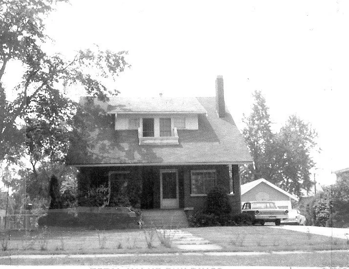 Black-and-white photo of a two-story bungalow with a covered front porch and dormer window; driveway to the right with a vintage station wagon and detached garage.