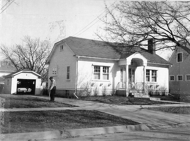 Black-and-white photo of a single-story bungalow with a porch and detached garage (car visible inside) and a man standing on the front lawn.