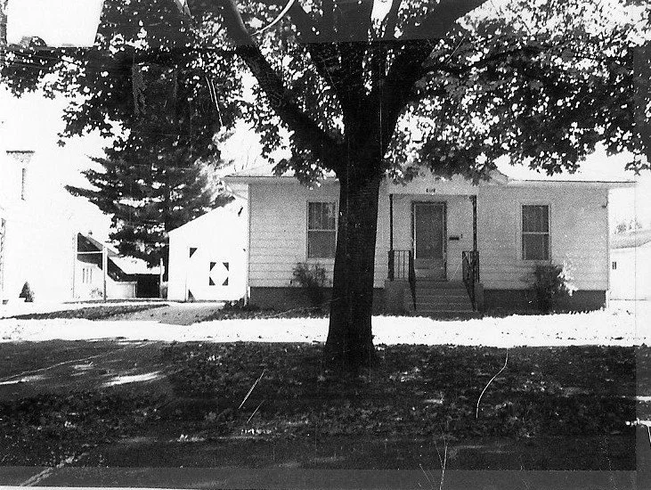 Black-and-white photo of a small single-story house with front steps and porch, a large tree centered in the foreground, and a garage to the left.