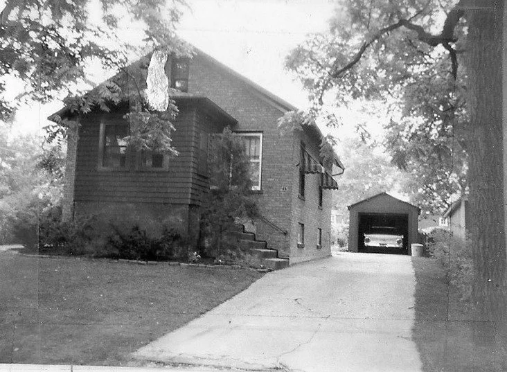 Black-and-white photo of a small brick bungalow with a front lawn and driveway leading to a detached garage where a car is parked.