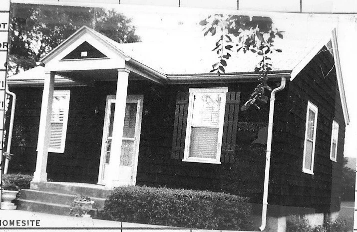 Black-and-white photo of a small one-story house with a gabled front porch, two columns, three windows with shutters, concrete steps and shrubs along the front.