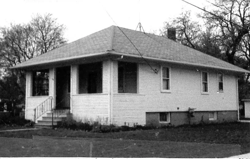 Black-and-white photo of a single-story bungalow with a covered front porch, steps, and a grassy yard.
