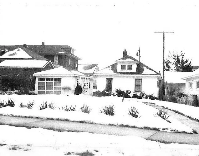 Black-and-white photo of a small single-story house with attached garage and snow-covered front yard on a residential street.