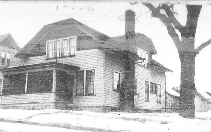 Black-and-white photo of a two-story wood-sided house with a covered front porch, tall chimney, a large bare tree, and a snow-covered yard.