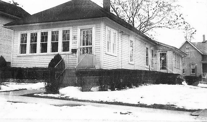 Snow-covered yard and sidewalk in front of a single-story white wood house with an enclosed front porch and steps up to the entrance.