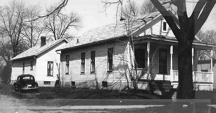 Black-and-white photo of two single-story wooden houses with a covered front porch, a large tree, and a parked vintage car.